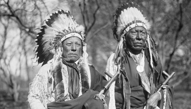 Two Native American men in traditional attire with feather headdresses and holding rifles.